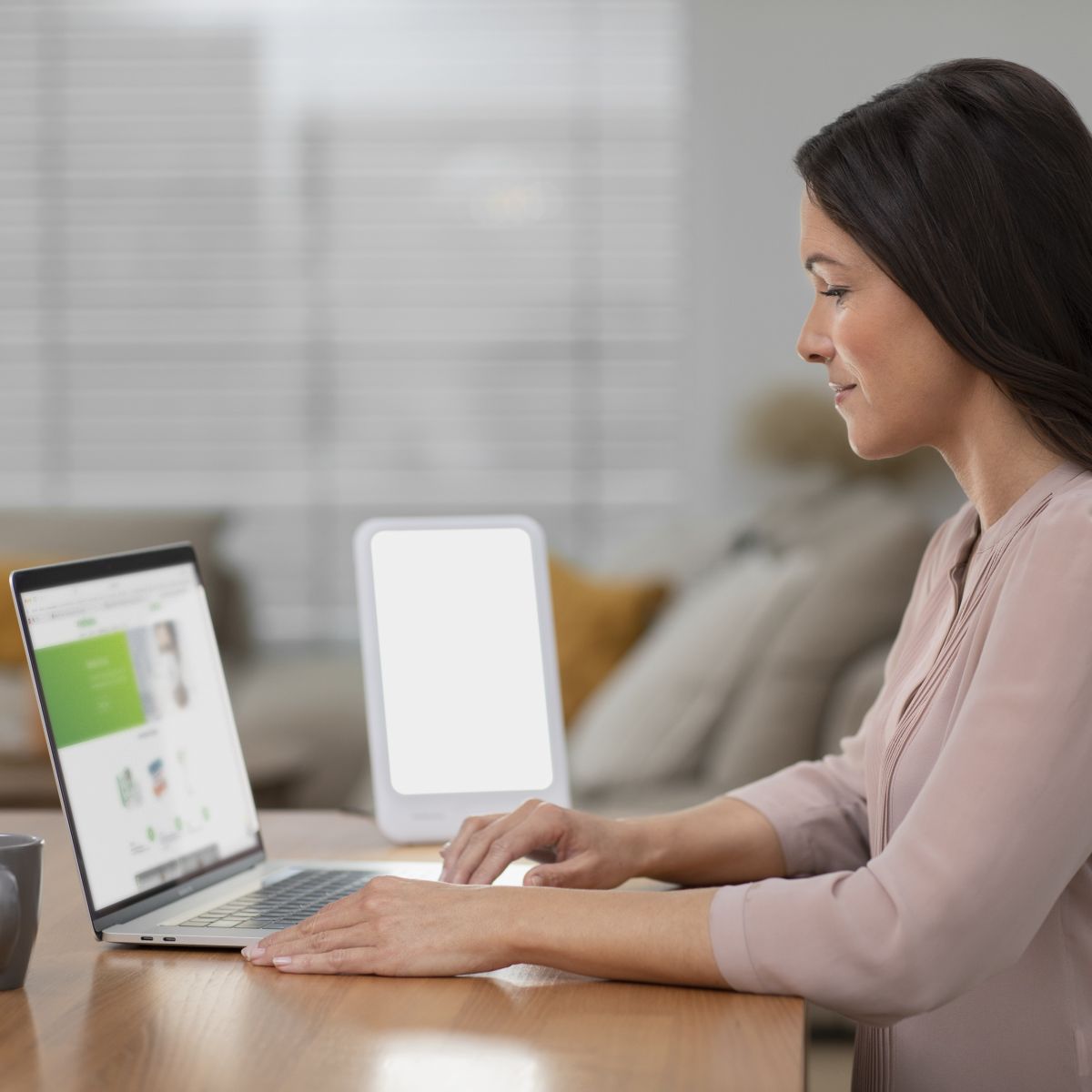 A woman sits at a table working on her laptop while a switched-on light therapy lamp provides bright, even illumination.